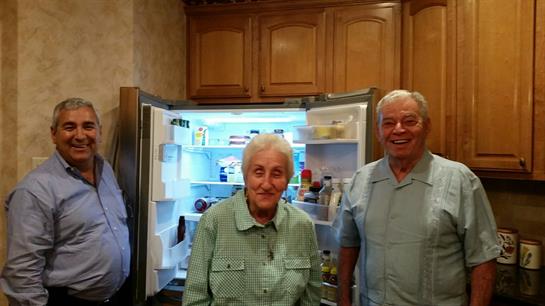 Three seniors share smiles and laughter together in a cozy kitchen with an open refrigerator.