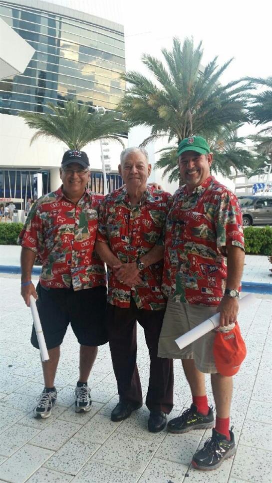 Three men wearing colorful shirts stand together outside, smiling in a lively tropical atmosphere.