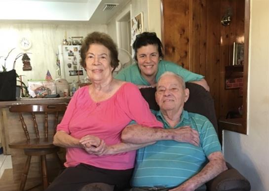 An elderly couple shares smiles with their caregiver in a warm, cozy living room.