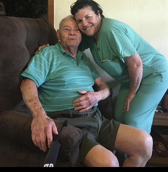 An elderly man sits in a cozy chair with a happy caregiver next to him, both smiling at the camera.