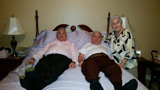 Three older individuals enjoy a moment of camaraderie while seated on a bed in a simple room.