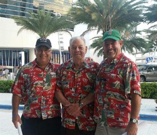 Three smiling men in vibrant tropical shirts stand together near a resort pool with palm trees.