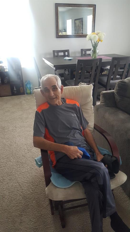 An older man sits comfortably in a chair in a sunlit living room, enjoying his time at home.