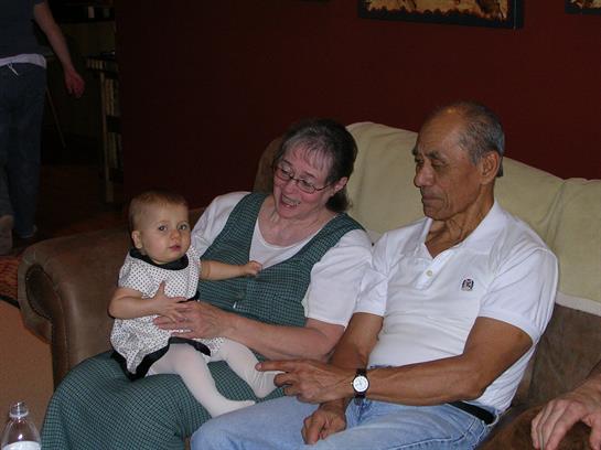 Grandparents smile while holding a baby in a warm living room setting with soft lighting.