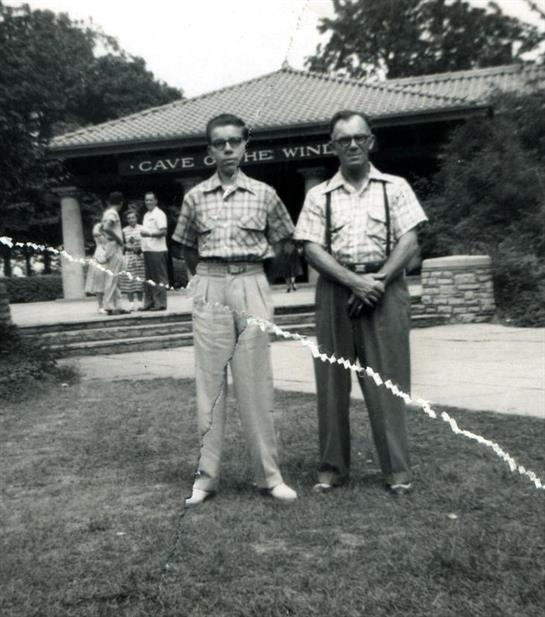 Two men pose confidently outdoors, with families enjoying the attraction in the background.