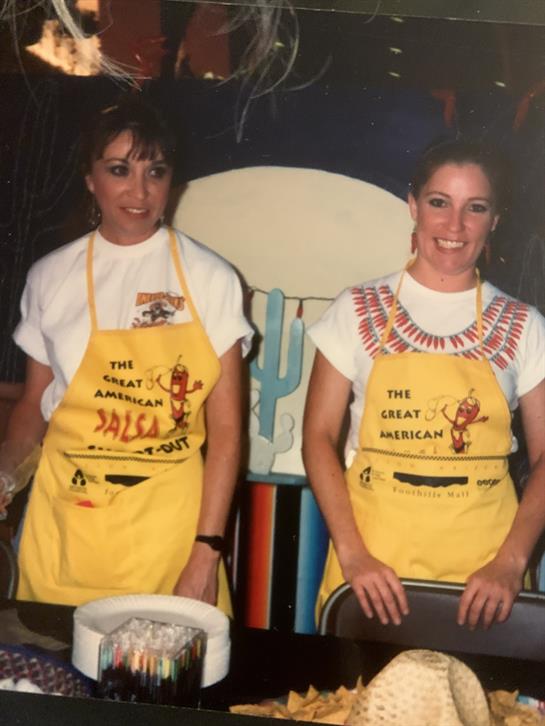 Two women in bright yellow aprons smile as they prepare food at a celebration.