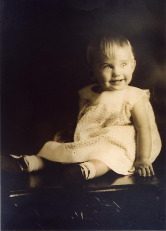 A cheerful toddler poses on a dark surface with a warm smile, dressed in a light dress.
