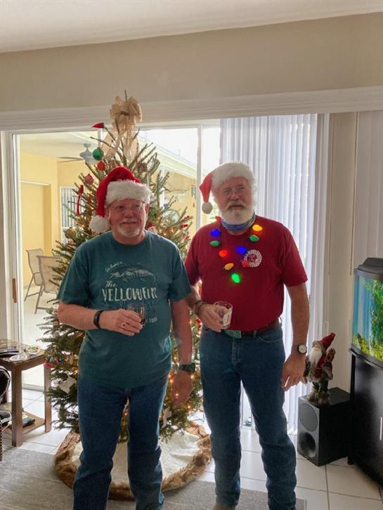 Two friends wearing Santa hats enjoy drinks in front of a decorated Christmas tree.