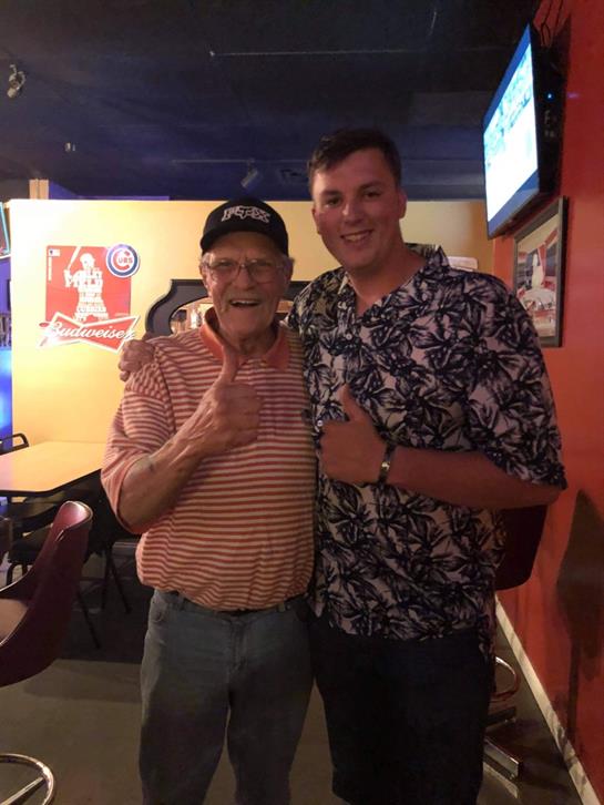 Two men pose together with thumbs up, showcasing smiles in a lively bar setting at night.