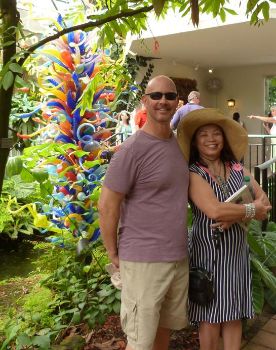 Couple stands joyfully next to an artistic glass installation surrounded by greenery.