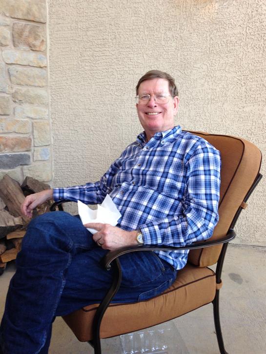 A man enjoys his time sitting in a chair, holding a paper while smiling in a sunny setting.