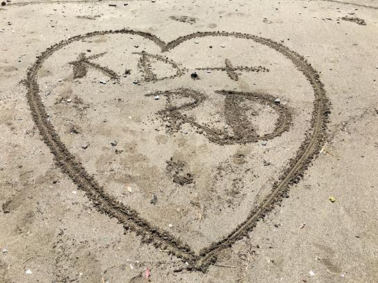 Heart symbol in sand features initials K.D. and R.D., celebrating love on a warm beach day.