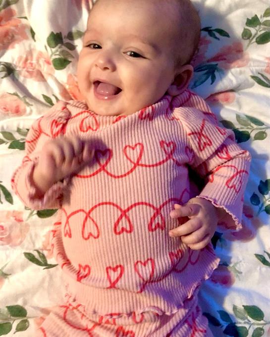 A joyful baby smiles while lying on a floral blanket, dressed in a charming red outfit with hearts.