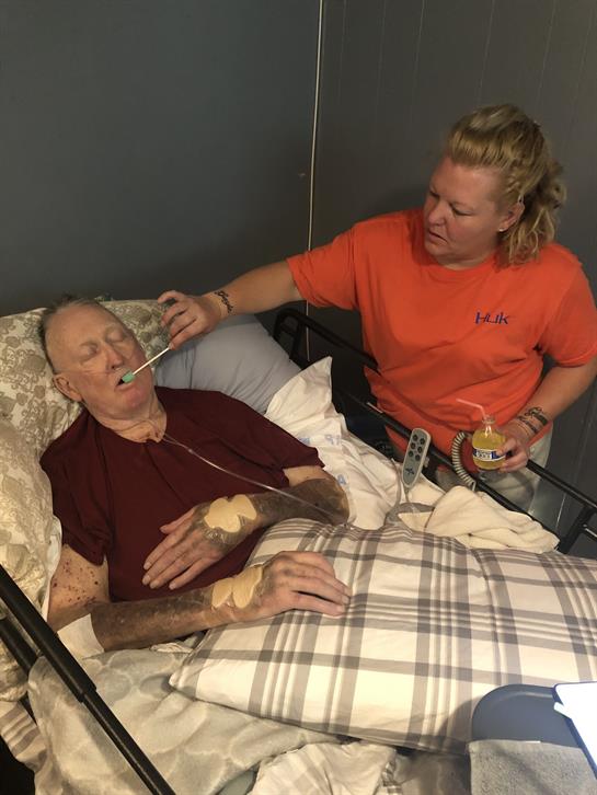 A caregiver gently helps an elderly man take his medicine while providing reassurance in bed.