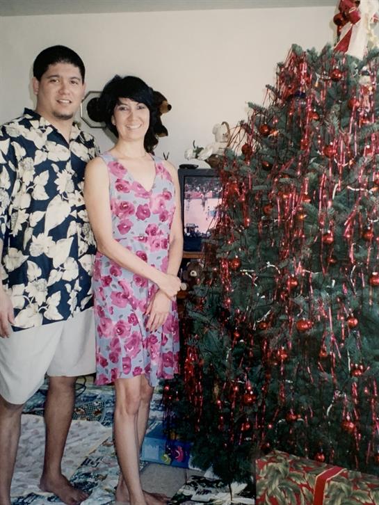 A couple stands together in a cozy living room, posing by a beautifully decorated Christmas tree.
