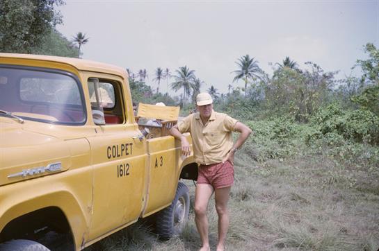 A man in shorts leans against a yellow truck in a lush, tropical environment.