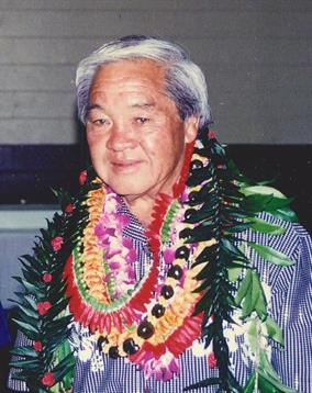 An elder in traditional attire wears vibrant leis, celebrating in a community hall.