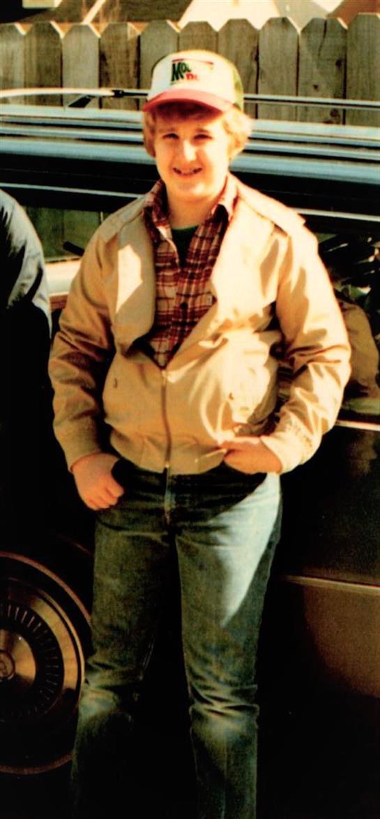 A young boy in a plaid shirt and jacket stands by a classic car, hands on hips.