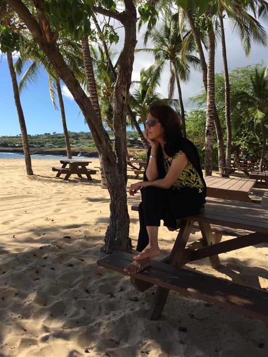 A woman relaxes on a wooden bench near the ocean, surrounded by swaying palm trees.