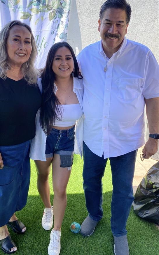Three people are standing close together, smiling in a sunny backyard with green grass.