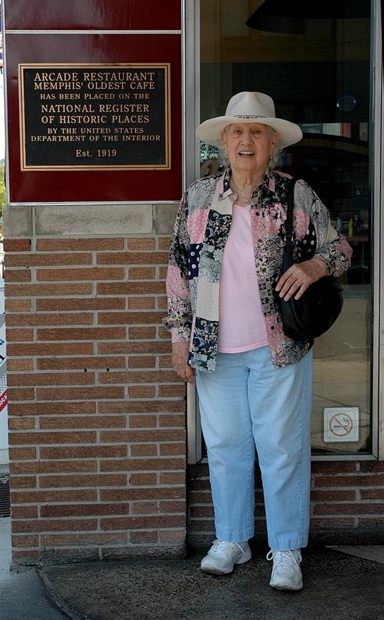 Older woman smiles while posing by a historic place sign, showcasing her stylish outfit and sun hat.