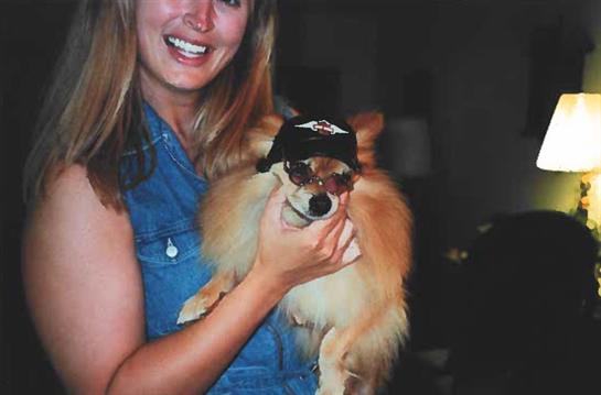 A woman happily poses with her fluffy dog in sunglasses and a hat, capturing a joyful moment.