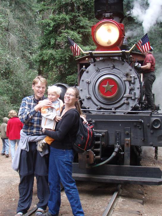 Family poses joyfully in front of a vintage steam train during a visit to a forest railway.