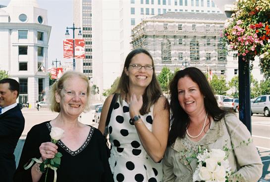 Three women enjoy a joyful moment together, holding flowers and smiling in a lively urban setting.