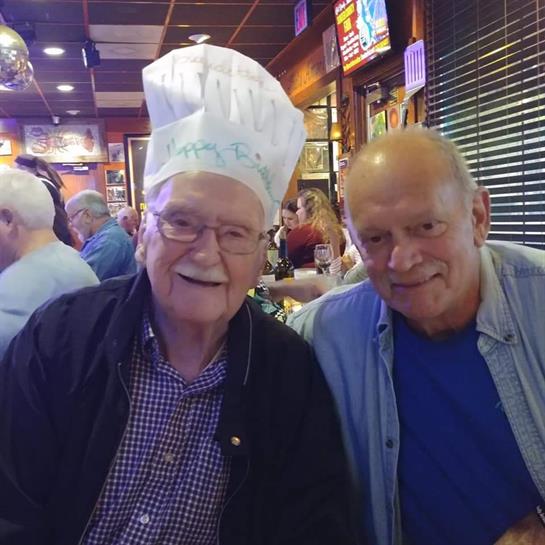 Two elderly men smile joyfully while seated at a busy restaurant filled with people.