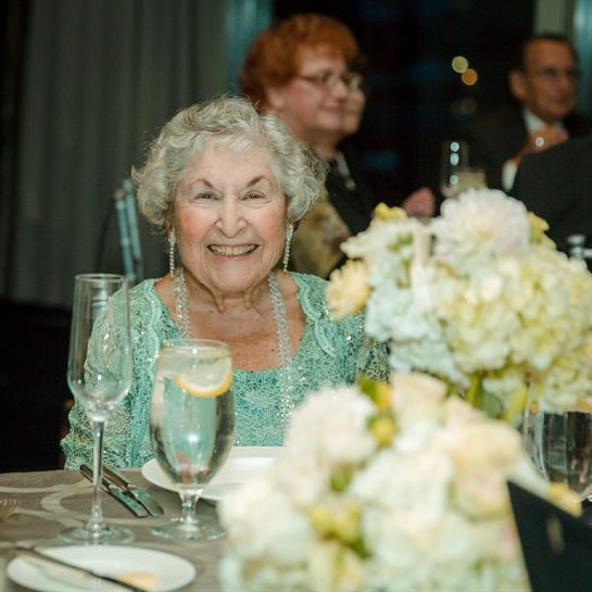 An elderly woman smiles brightly at a formal dinner, surrounded by friends and elegant floral decor.