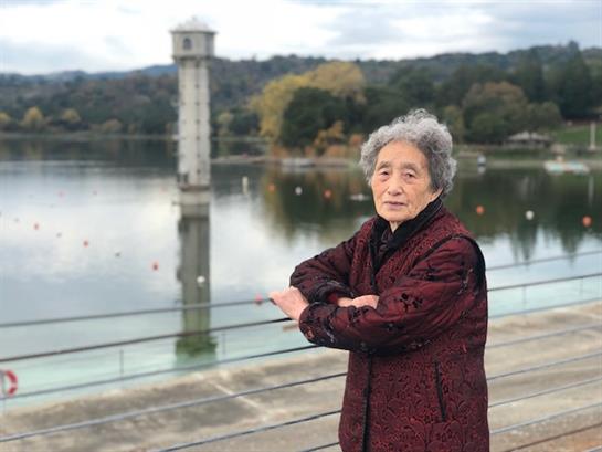 An elderly woman stands with arms crossed by a serene lake reflecting trees and a tower.