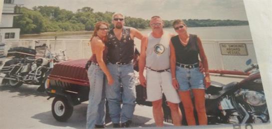 Four friends pose together by a river, smiling and dressed casually, enjoying a summer day.