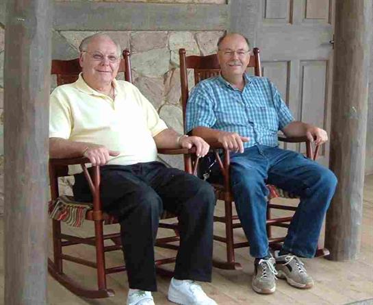 Two older gentlemen sit comfortably in rocking chairs, enjoying each other's company indoors.