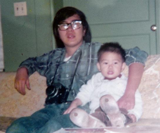 A father sits on a couch with his young child, both smiling and enjoying a relaxed moment together.