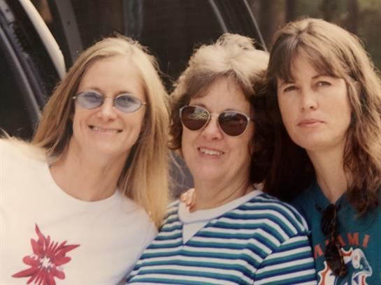 Three women smile and pose together on a sunny day, enjoying each other's company outdoors.