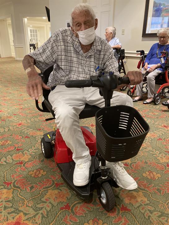 An elderly man on a mobility scooter enjoys an indoor gathering at a retirement community.