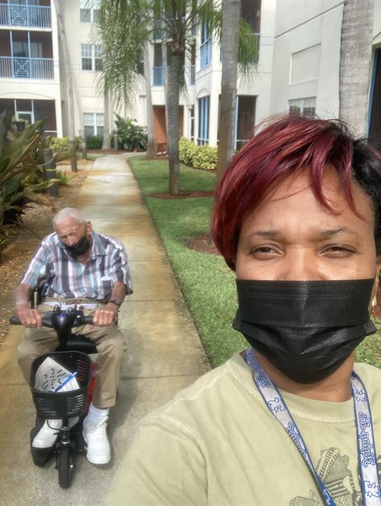 A caregiver helps an elderly man on a scooter while walking through a residential area.