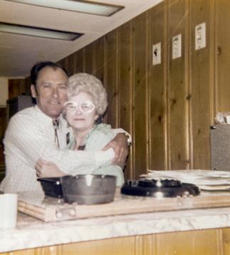 A couple embraces affectionately in a warm kitchen, surrounded by cooking utensils and lively decor.