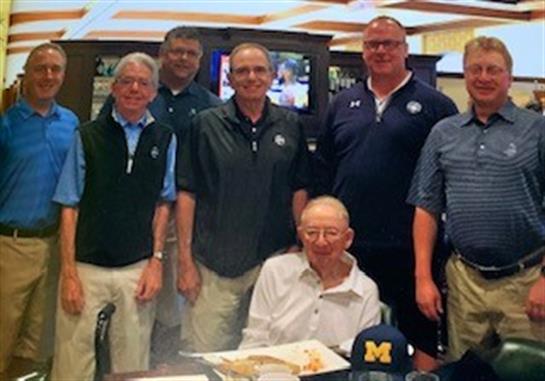 Seven friends stand together while one sits at a table, all smiling and enjoying the moment.