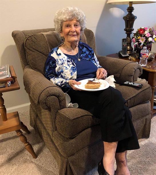 Elderly woman sits in a comfy chair, smiling while holding a plate of food in a cozy living room.