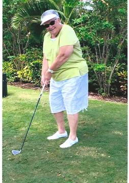 An elderly woman in casual attire swings a golf club on a green lawn, surrounded by plants.