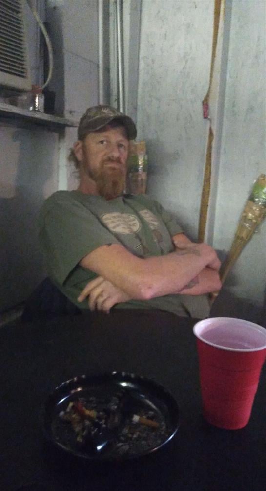 A man sits comfortably indoors, wearing a green shirt and hat, looking contemplative while relaxing.