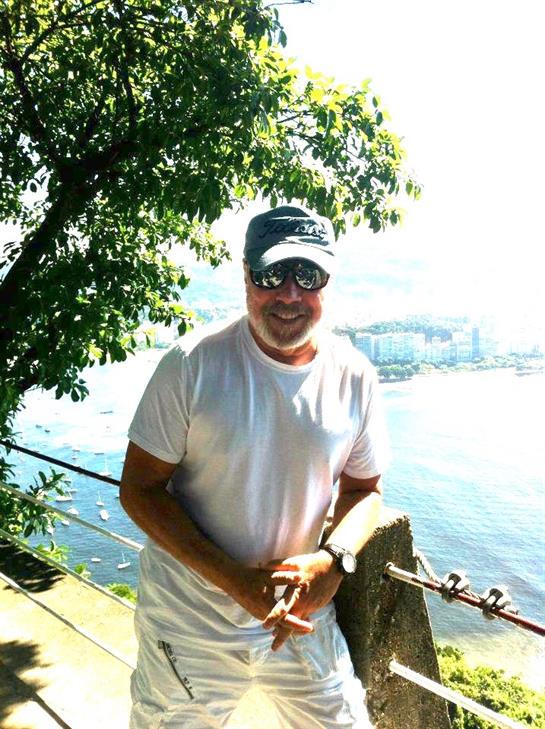 A man in a white shirt stands near a waterfront, smiling against a sunny backdrop with trees.
