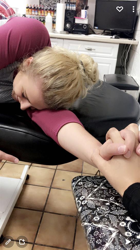 A woman leans in, gripping her friend's hand tightly, showing her determination during a tattoo.