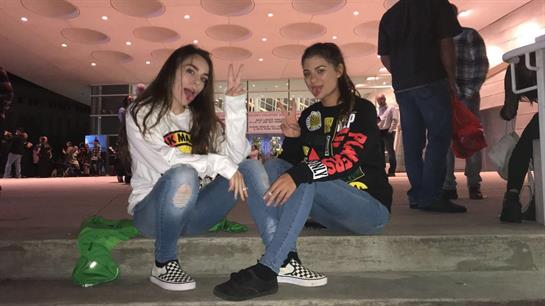 Two young women sit on steps, smiling and making peace signs in a vibrant urban atmosphere.