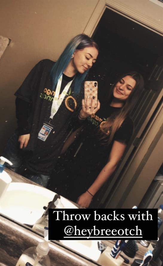 Two friends smiling together in a bathroom mirror, showcasing colorful hair and event attire.