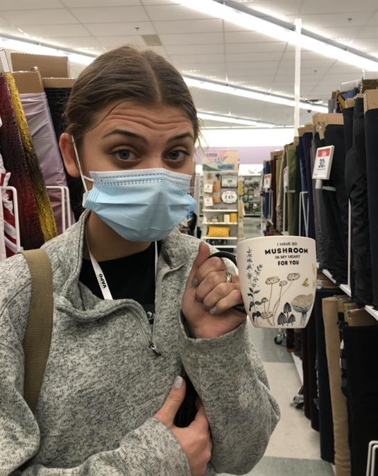Young woman with a mask points at a coffee cup while browsing fabrics in a store.
