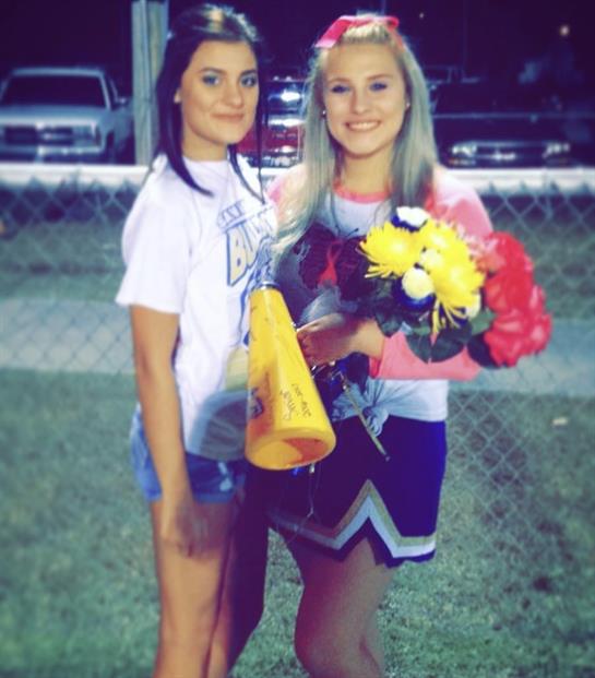 Two cheerleaders wearing uniforms and casual clothes pose with flowers and a megaphone after a game.