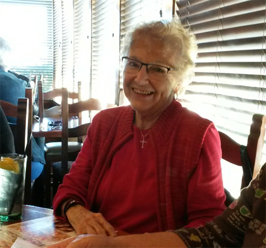 An elderly woman with glasses is smiling warmly while seated at a table in a restaurant.