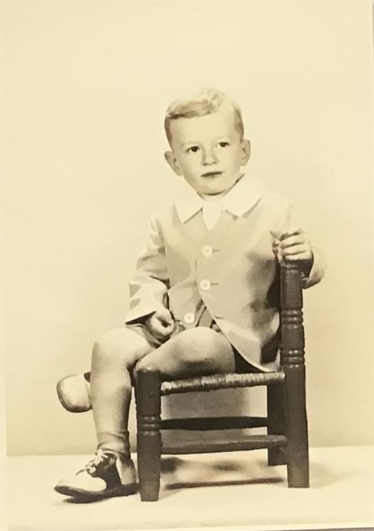 A young boy dressed in a light suit sits on a small wooden chair, giving a charming expression.